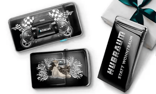 Drei schwarze Geldbörsen mit Auto-Designs und karierten Flaggen, vor einem weißen Hintergrund. Eine zeigt ein Auto mit Rädern, eine andere ein Paar, und eine trägt den Text „Hubraum statt Wohntraum“.