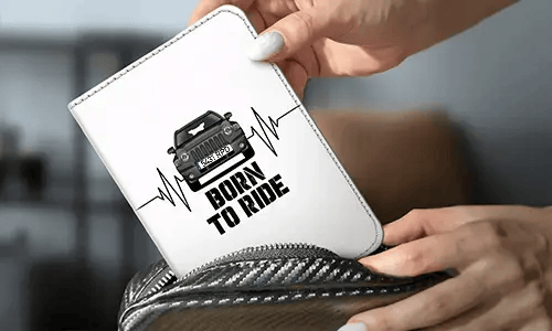 Personne tient un porte-monnaie blanc avec voiture noire et design battements de cœur, texte BORN TO RIDE, sac en fibre de carbone en arrière-plan