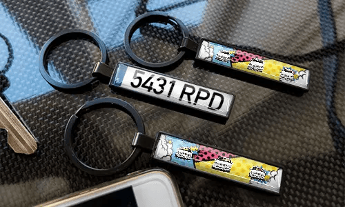 Comic-style license plate keychains displayed on a carbon-textured surface with colorful car designs