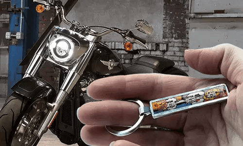 Metalowy brelok w stylu komiksowym z ilustracją motocykla, trzymany w dłoni obok motocykla