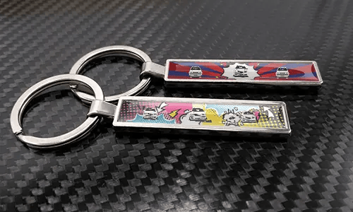 Comic-style license plate keychains with car graphics placed on a carbon-textured surface