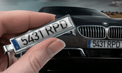 Sarjakuvatyylinen rekisterikilpien avaimenperä, jossa mukautettu numero, pidettynä kädessä lähellä vastaavaa autokilpeä